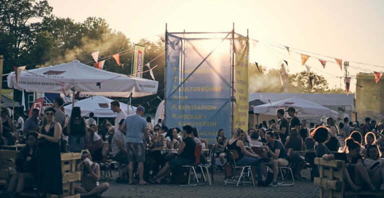 Outdoor festival scene with people socializing at wooden tables, tents with branding, and flags. Sun setting in the background creating a hazy, golden atmosphere.