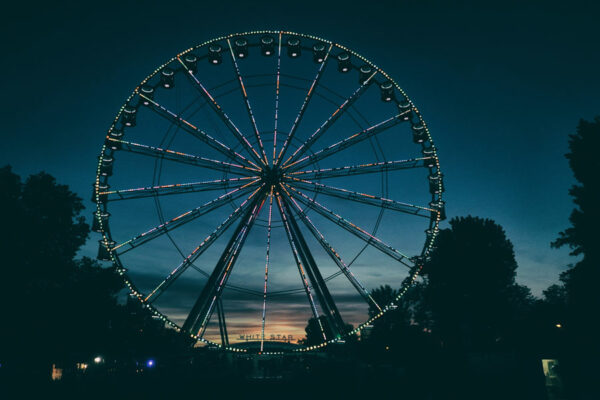 A Ferris wheel illuminated with white and colored lights against a dusky sky, with trees silhouetted in the foreground.