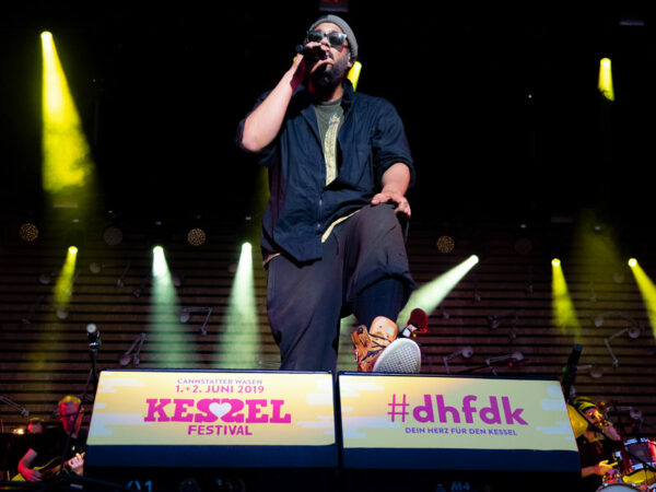 A musician wearing sunglasses and a cap sits on the edge of the stage, performing into a microphone at the Kessel Festival, with spotlights and the festival's banner visible in the background.