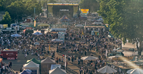 A crowded outdoor music festival with multiple vendor stalls, a large stage in the background, and many attendees spread out across the venue.