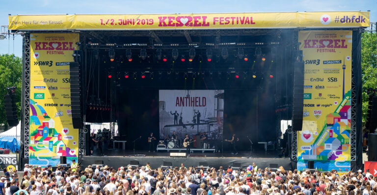 A music festival scene with a crowded audience in front of a large stage displaying the word 'ANTHELD' on the backdrop, with band members performing. Banners on either side of the stage announce the event as 