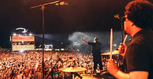 A vibrant concert scene from the perspective of the stage, featuring a drummer in the foreground on the right and a vocalist engaging a large crowd of fans in the background at night, with festival lighting and a 