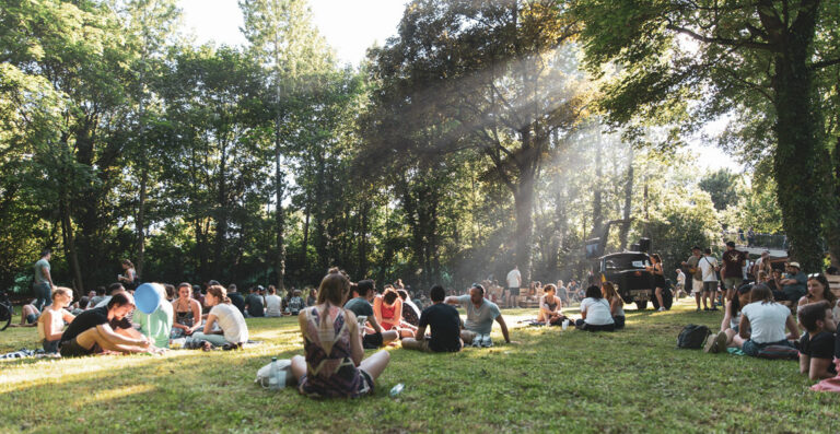 A group of people sitting and lounging on the grass in a sunlit park, with trees and a food truck in the background. Rays of sunlight are streaming through the foliage.