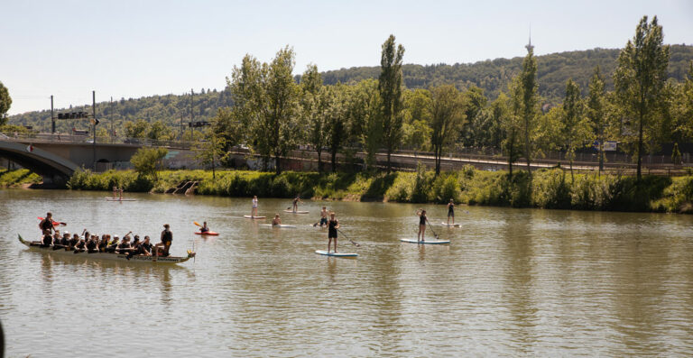 A group of people paddling a large canoe and others stand-up paddleboarding on a calm river, with a bridge and greenery in the background on a sunny day.
