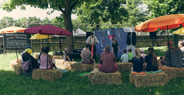 An outdoor puppet theater performance with a small audience sitting on hay bales under colorful umbrellas, with a chalkboard event schedule on the left.