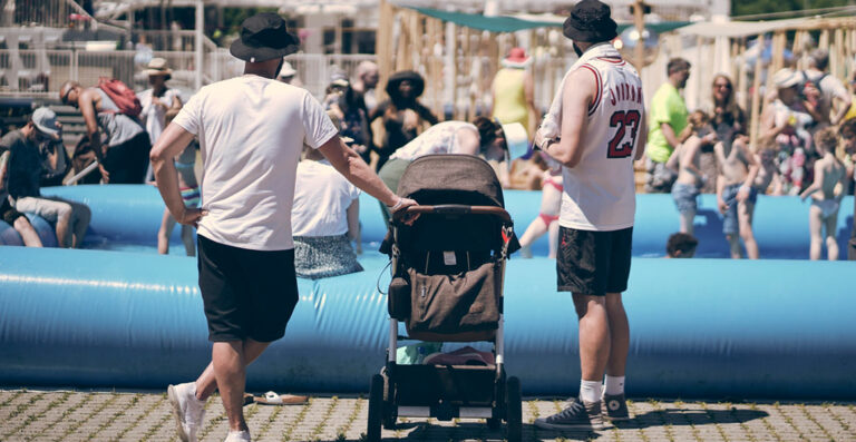 Two men standing next to a baby stroller at an outdoor event with people relaxing in the background.