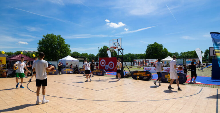 Outdoor basketball event with players practicing on courts amid sponsor banners and tents, under a clear blue sky.