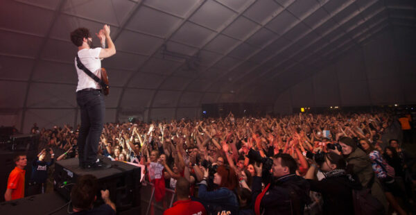 A musician stands on a speaker with his back to the camera, raising his arms to an enthusiastic crowd inside a large tent at a concert.