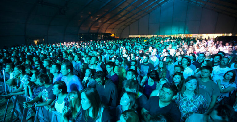 A large crowd of people inside a tent at a concert or event, illuminated by green and blue stage lights.