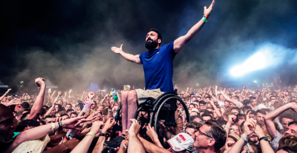 A man in a wheelchair is raised above a cheering festival crowd, arms outstretched with excitement.