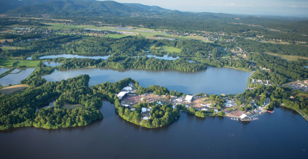 Aerial view of a large outdoor festival set up on the shores of a lake with multiple stages, tents, and a crowd, surrounded by lush greenery, with mountains in the background.