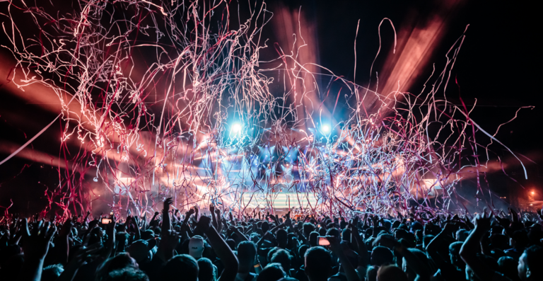 A lively concert crowd with hands raised, silhouetted against a stage with bright lights and dynamic streams of confetti in the air.