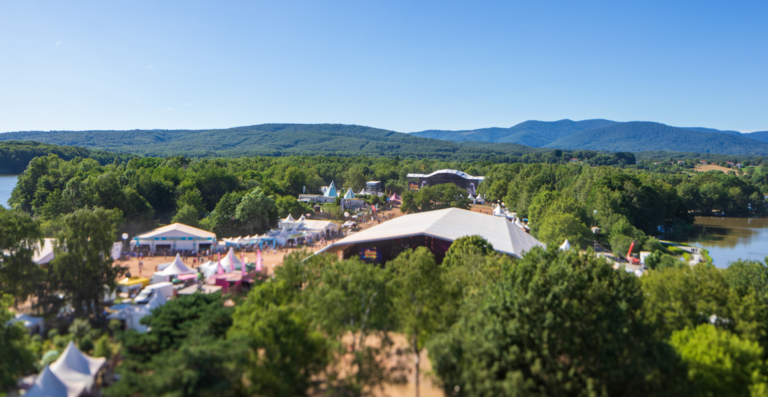 An aerial view of an outdoor festival by a river with tents, pavilions, and surrounding green hills under a blue sky.