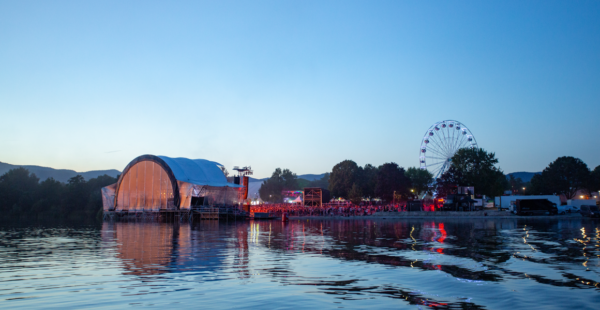 An outdoor festival by a lake at dusk, featuring a large stage covered with a transparent tent, a Ferris wheel, and a crowd of people, with lights reflecting on the water.