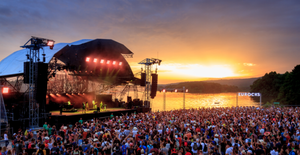 A large outdoor music festival with a vibrant sunset in the background, featuring a crowded audience in front of a stage with performers and bright stage lights.