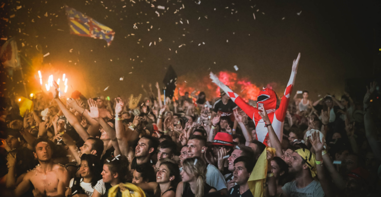 Crowd of enthusiastic concert-goers cheering with hands raised, some holding flares, in a vibrant atmosphere with confetti in the air.