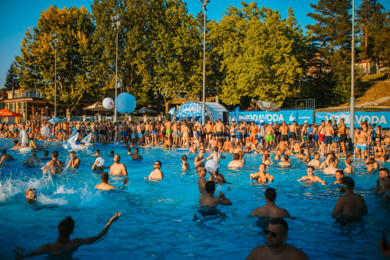 A crowded outdoor pool with people swimming, talking, and playing with inflatable balls, with trees and tents in the background on a sunny day.