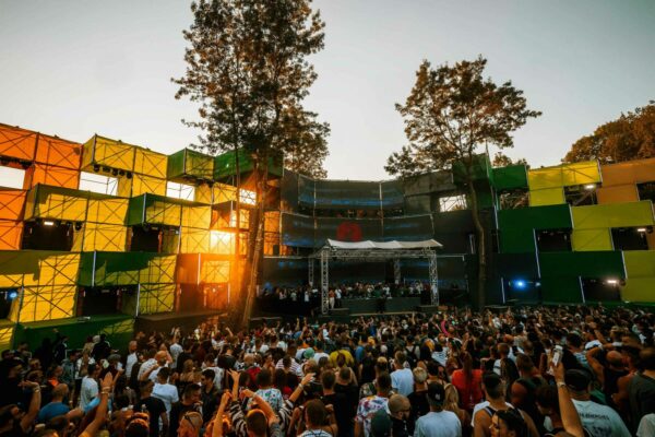 A crowd enjoying an outdoor music event at sunset, with a DJ on stage and colorful stage design.