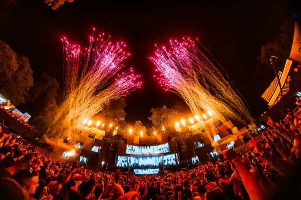 A vibrant outdoor music festival scene at night with a large crowd of people enjoying the performance under a display of red fireworks exploding above the stage.