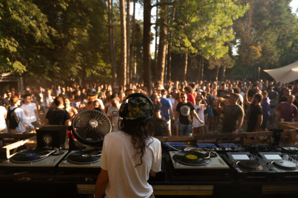 A DJ facing a crowd at an outdoor music event, with turntables in the foreground and trees in the background.