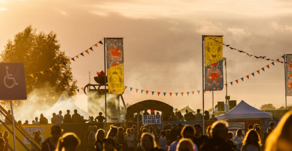 Outdoor festival at sunset with people gathered around stalls and stages, decorative banners and bunting in the foreground, against a backdrop of a hazy sky.