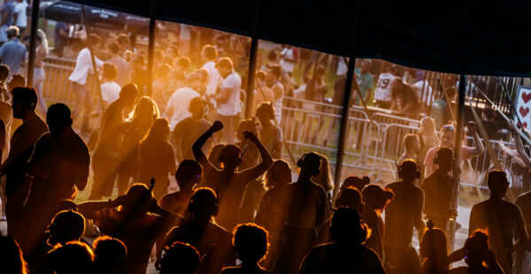Silhouettes of people at an outdoor music festival with sunset lighting, dancing and enjoying themselves under a tent.