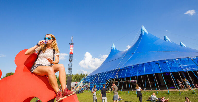 A young woman sitting on a large red puzzle piece sculpture is drinking from a can, with a blue circus tent and people enjoying a sunny day at an outdoor festival in the background.