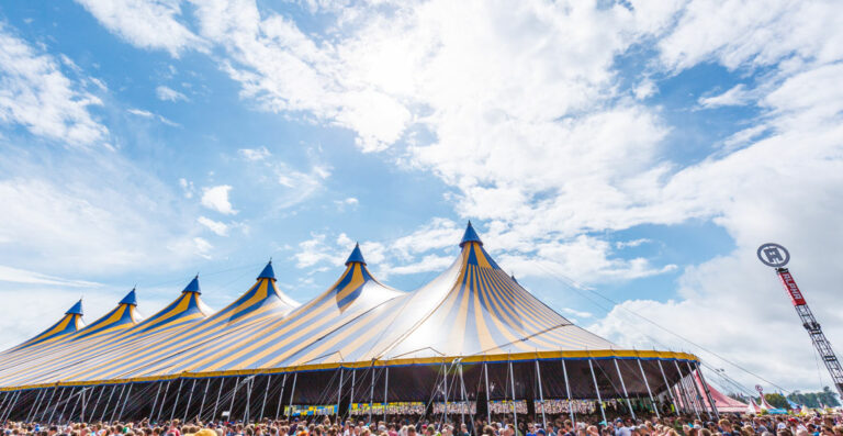 A large crowd gathers outside a massive blue and yellow striped circus tent under a blue sky with scattered clouds.