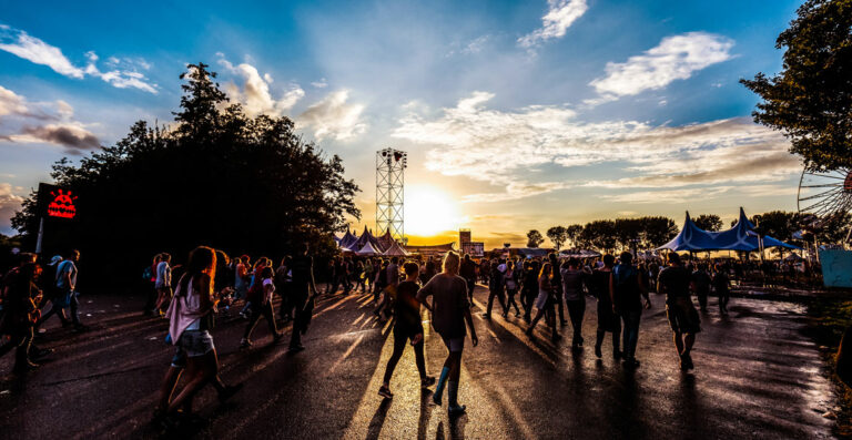 People walking at an outdoor festival during sunset with tents, trees, and a clear sky in the background.