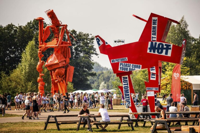 An outdoor festival scene with a large orange sculpture resembling a robot on the left and an oversized red sculpture of a pilot's head and a hand with various phrases in white lettering on the right. People are walking around and sitting at picnic tables in the foreground.
