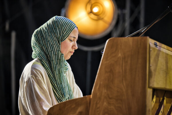A woman wearing a hijab is standing at a wooden lectern, looking down, possibly reading or speaking, with a warm light glowing behind her.