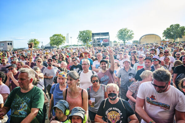 A large crowd of people at an outdoor festival with some individuals wearing sunglasses, hats, and one person with a face mask, amidst a sunny day with a clear blue sky.