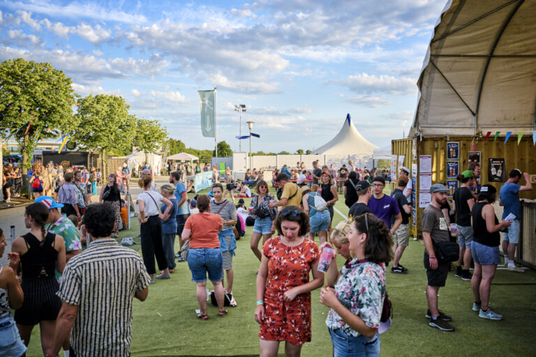 A lively outdoor festival scene with people mingling, some walking and talking, with tents and flags in the background under a clear blue sky.