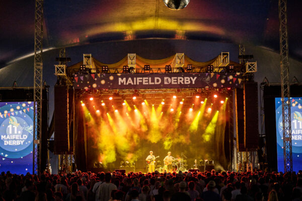 A concert scene at the Maifeld Derby festival with a crowd of spectators watching a band perform on a stage with colorful lighting and large screens displaying the event's logo.