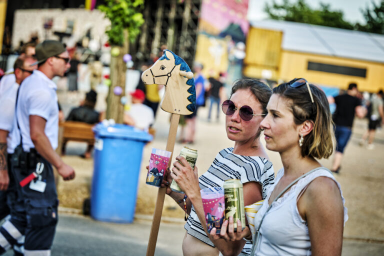 Two women holding cans and talking at an outdoor festival with a wooden hobby horse in the foreground and blurred festival-goers in the background.