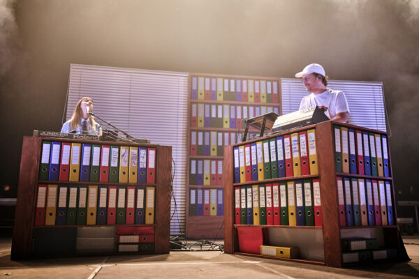 Two musicians performing on stage behind custom synthesizer stands designed to resemble oversized office file binders, with a colorful backdrop of more file binders.