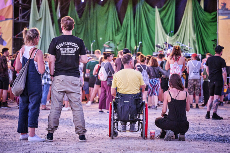 A diverse crowd of people at an outdoor concert, with some standing and others sitting on the ground or in wheelchairs, facing a stage with green curtains and a drum set in the background.