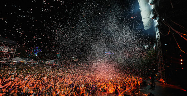A lively outdoor concert with a dense crowd of cheering people and confetti raining down from above.