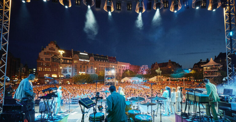 View from a stage at an outdoor concert showing musicians facing a large crowd with buildings and a Ferris wheel in the background during evening hours, with stage lights casting beams into the sky.