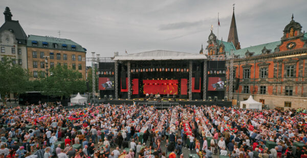 A large outdoor concert with a crowded audience in a city square. The stage features red accents and large screens displaying performers, set against a backdrop of striking historical buildings under a cloudy sky.