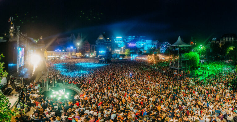 A panoramic view of a crowded outdoor music festival at night with a stage brightly lit on the left, dense audience in the center, and city buildings illuminated in the background.