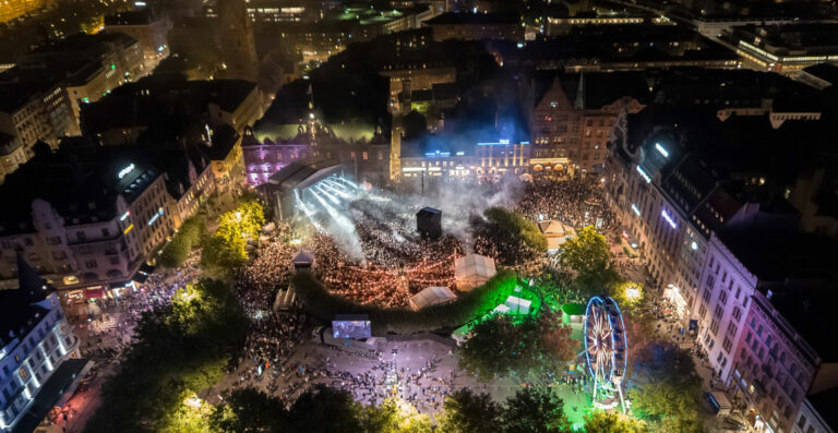 Aerial night view of a crowded outdoor concert with stage lights, surrounded by city buildings and a Ferris wheel, seen from above.