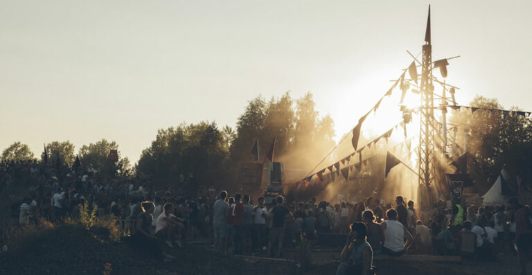 A large crowd of people gathered at an outdoor festival during sunset, with a dusty atmosphere and a large, makeshift wooden structure in the background adorned with flags.