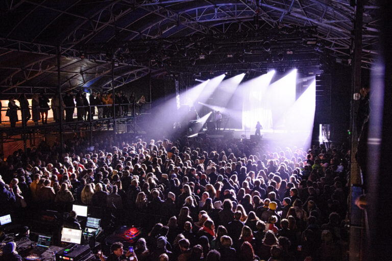 A lively concert scene with a large crowd watching a performance under bright stage lights, with some attendees on an elevated platform to the left. Sound equipment is visible in the foreground.