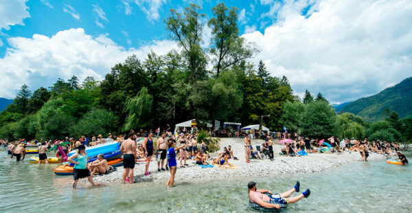 A busy riverbank with people in swimwear relaxing, sunbathing, and floating on inflatable rafts on a sunny day, surrounded by green trees and hills under a partly cloudy sky.