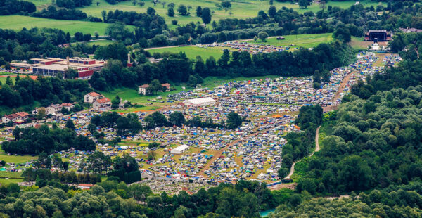 Aerial view of a large outdoor festival with tightly packed tents and vehicles amidst green trees and open grassy areas.