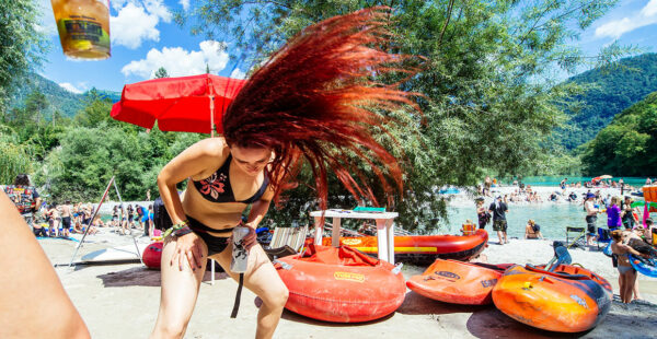 A woman with long, flowing red hair flips her head forward next to a river where people are swimming and lounging on a sunny day, with kayaks and a red umbrella in the foreground.