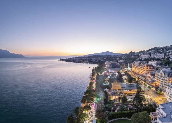 A panoramic twilight view of a coastal city with illuminated streets and buildings beside a calm sea, with hills in the background under a pastel sky.