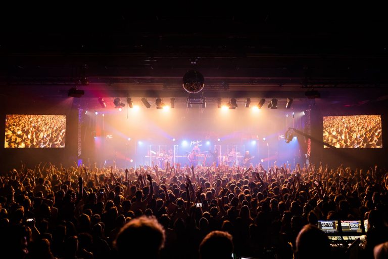 A crowded concert hall with a live band performing on stage, bright stage lights, and two large screens displaying the audience; fans are cheering with their hands up in the air.