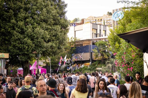 A bustling outdoor event with a diverse crowd of people walking and socializing. Colorful signage and festival decorations are visible amongst the trees and modern buildings in the background.